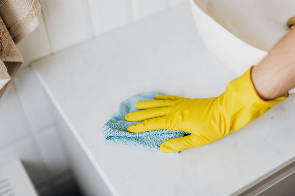 From above crop unrecognizable person with microfiber cloth wearing yellow rubber glove and cleaning white marble tabletop of vanity table with washbasin in bathroom